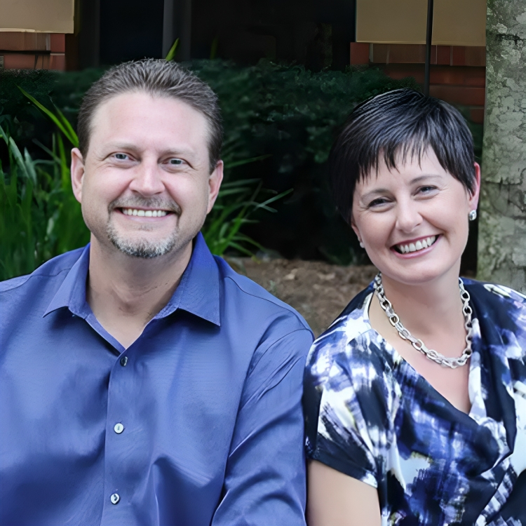 Two smiling dental professionals pose together outdoors in front of greenery.
