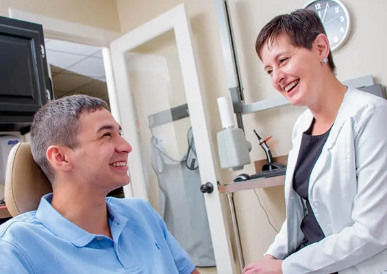 Dentist in white coat smiling at male patient in blue shirt in modern dental office treatment room.