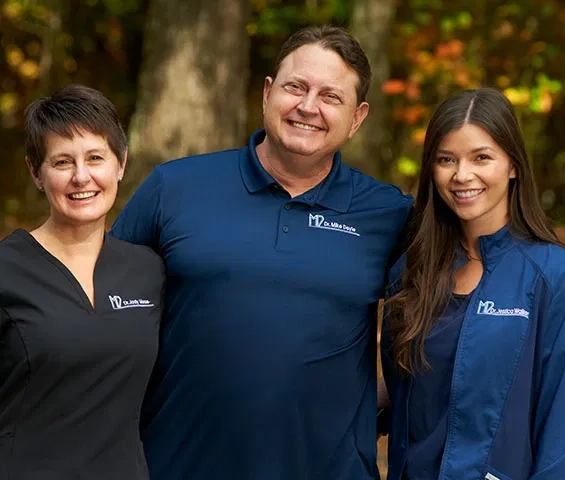 Three dental professionals in branded scrubs smiling outdoors with blurred green foliage background.