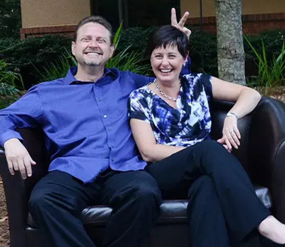 Two smiling adults sitting together on a dark couch outdoors, relaxed and friendly pose.
