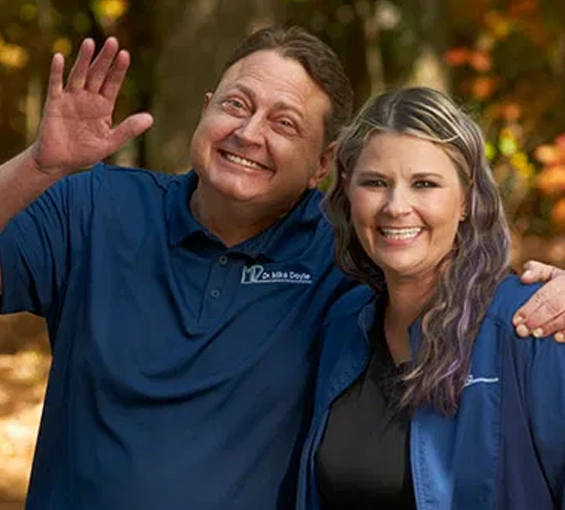 Two dental professionals in blue scrubs smiling and waving in front of holiday decorations.