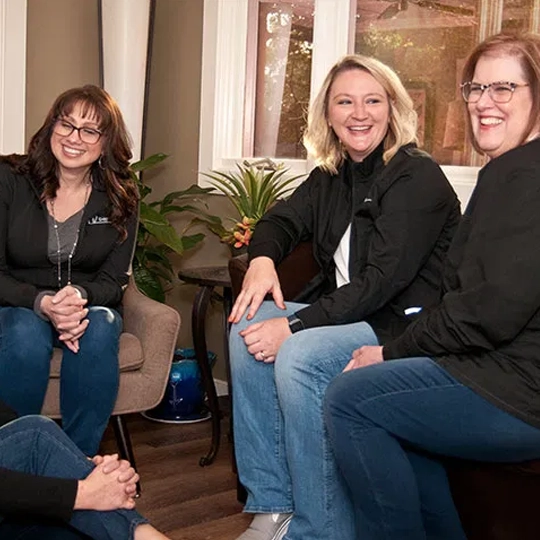 Three women smiling at camera in professional dental office setting with plant decor.
