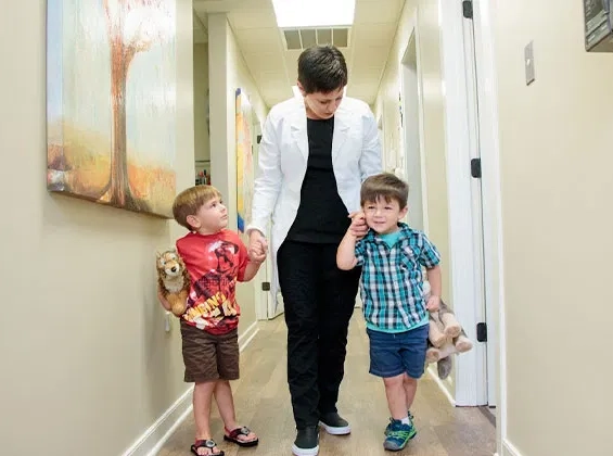 Pediatric dentist walks with two young children down bright dental office hallway.