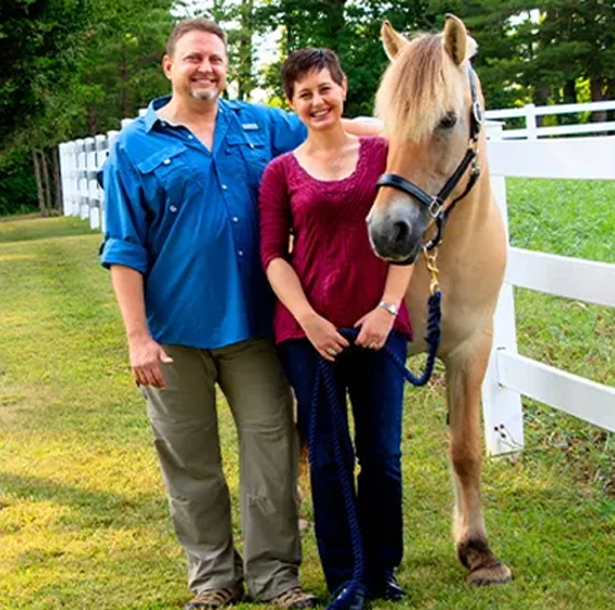 Two people standing with a horse on a grassy property with white fencing.